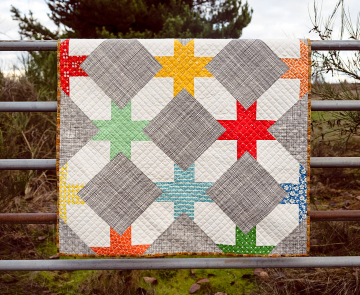 Bright and colorful baby quilt inspired by vintage children’s books, featuring a mix of red, green, yellow, and vivid blue fabrics with a crosshatch quilting design and ruler-print binding.